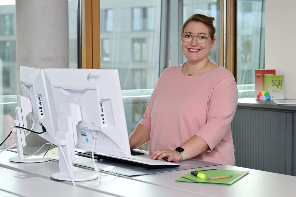 Eine freundliche Mitarbeiterin steht am Schreibtisch.