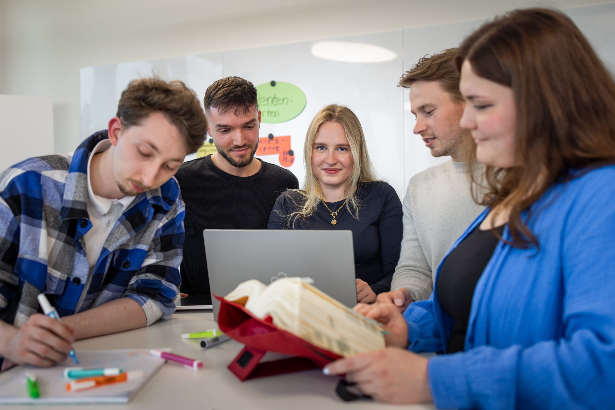 Auszubildende stehen an einem Tisch. Ein Auszubildender schreibt, drei schauen auf einen Laptopmonitor, eine liest im Sozialgesetzbuch