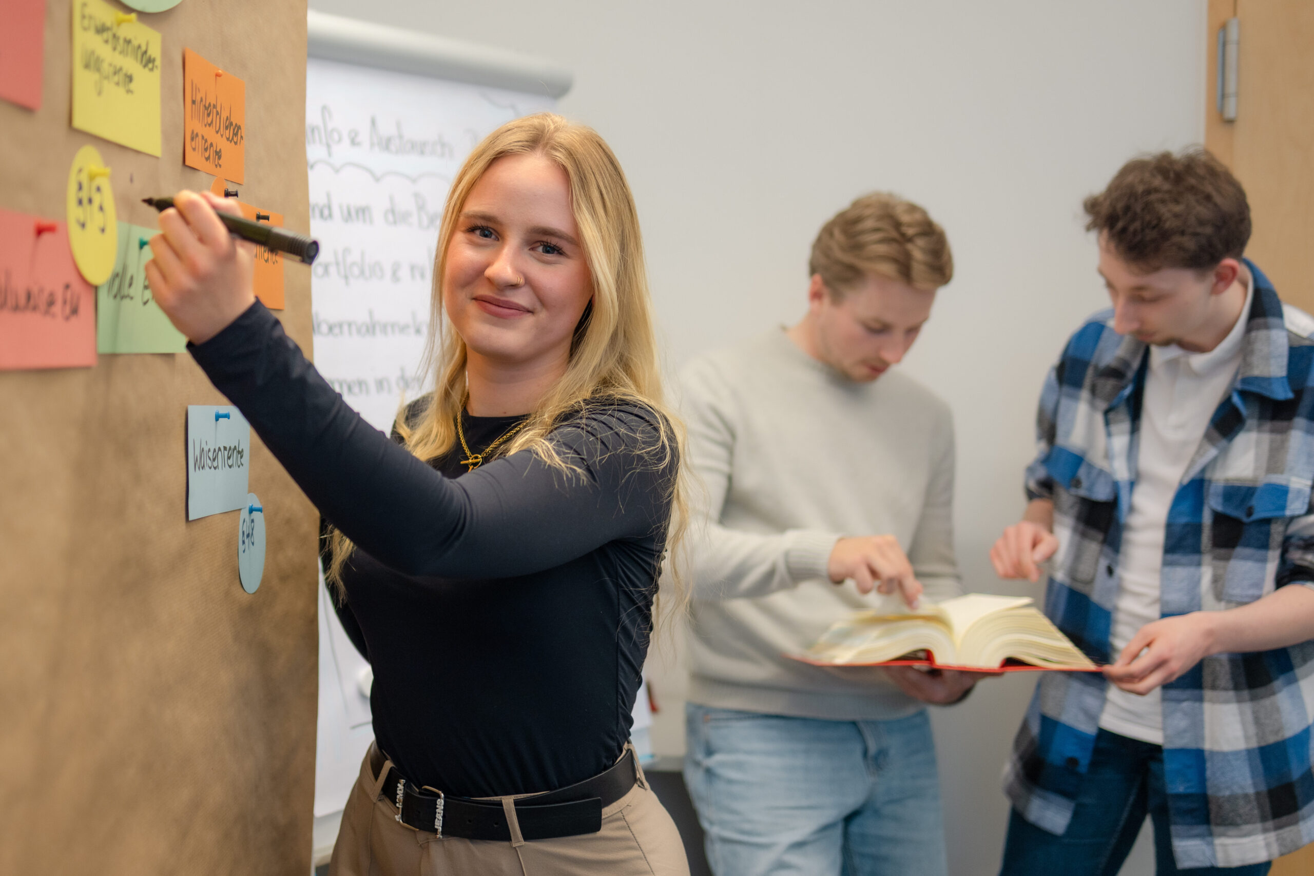 Auszubildende schreibt an Metaplanwand, im Hintergrund zwei weitere Auszubildende mit Sozialgesetzbuch in der Hand